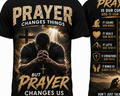 Black short-sleeve graphic T-shirt shown front and back. The front features large distressed gold text reading 'PRAYER CHANGES THINGS BUT PRAYER CHANGES US' with a central praying man, cross, and light rays.